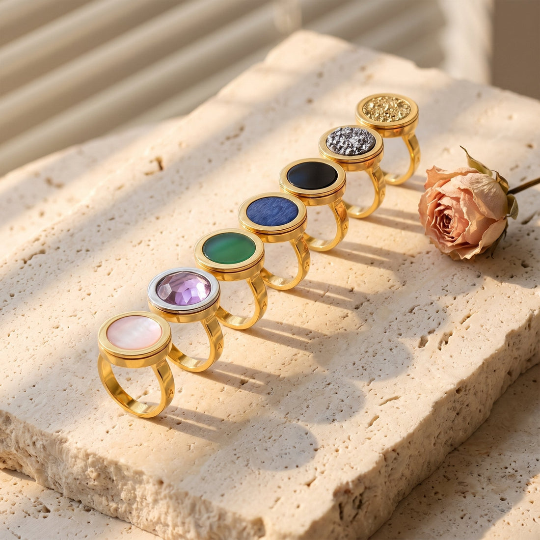 Sete anéis Eterno da Aimer em fila sobre travertino ao sol, cada um com uma pedra: madrepérola, ametista, ágata verde, feldspato azul anil, obsidiana, drusa platina e drusa ouro